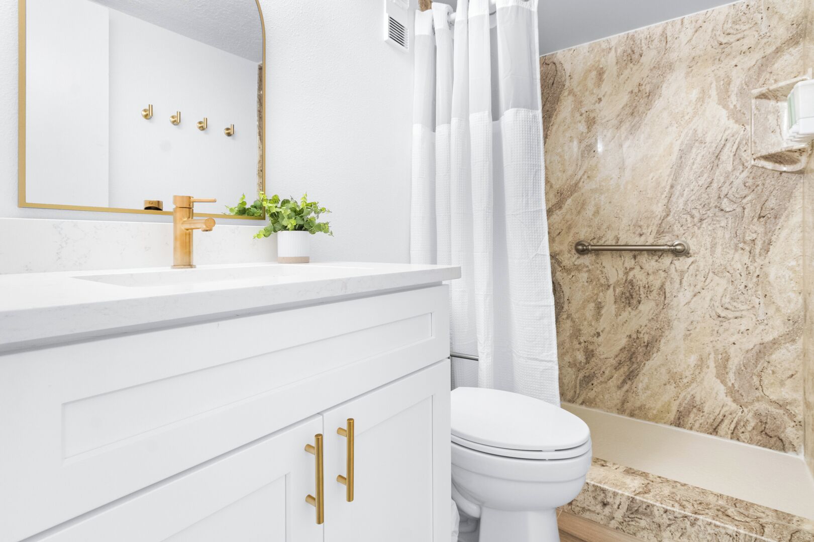 A bathroom with a white vanity, gold fixtures, a white toilet, and a marble shower wall.