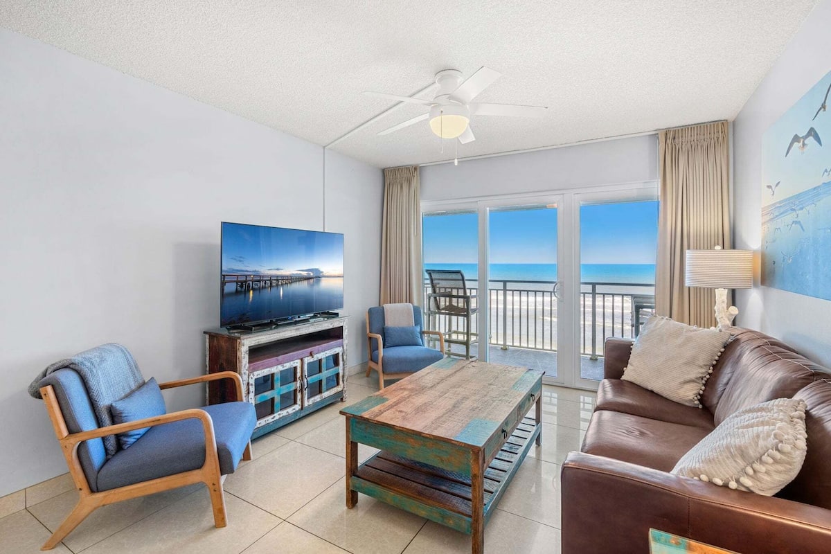 A living room with a TV, wooden coffee table, leather sofa, and a balcony with a view of the sea.