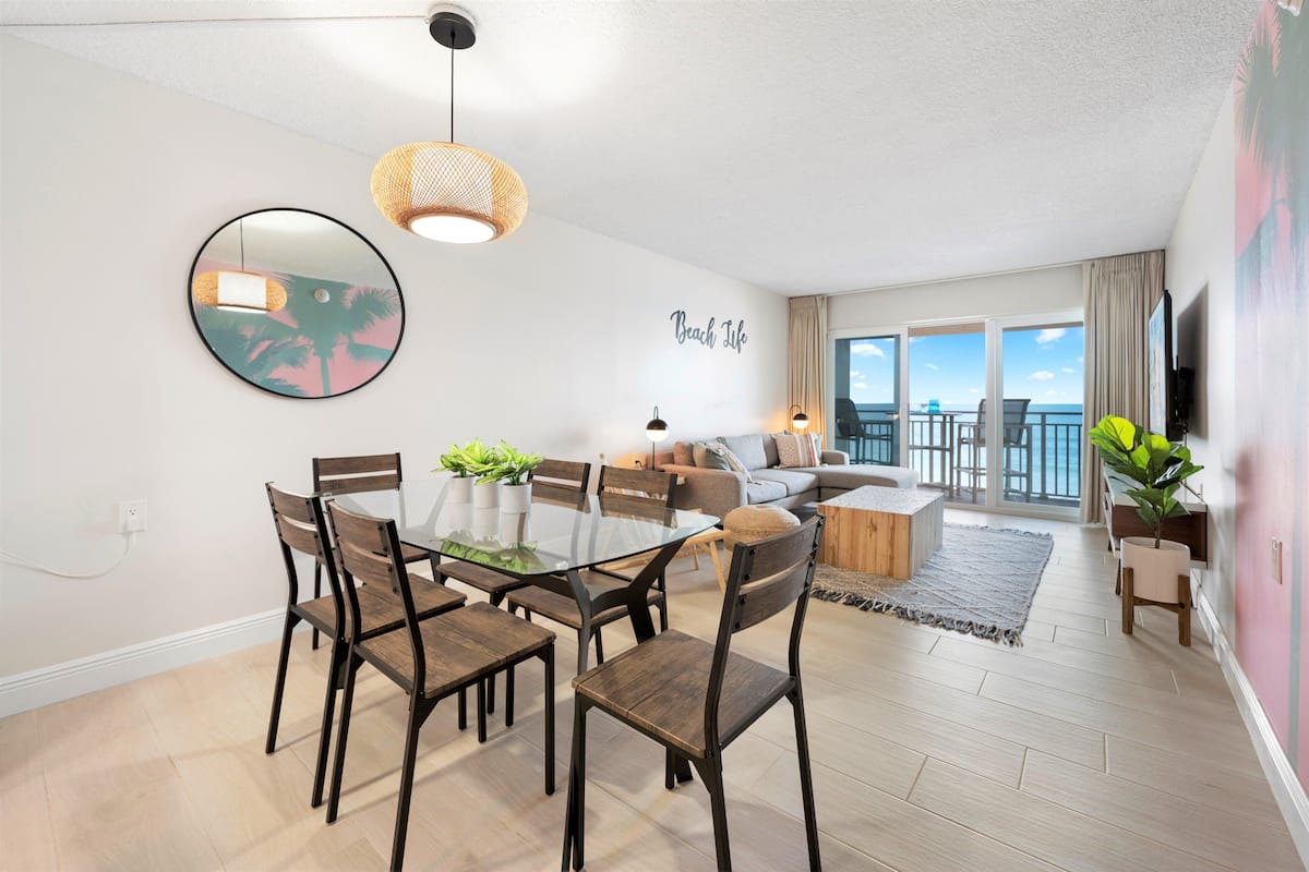 A modern living room with a glass dining table, wooden chairs, a sofa, and a balcony with a view.