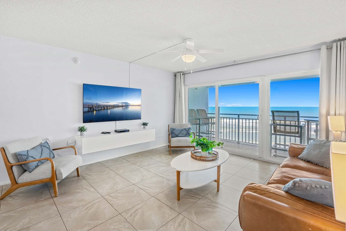 A modern living room with a TV, seating, a coffee table, and a balcony with a view of the sea.