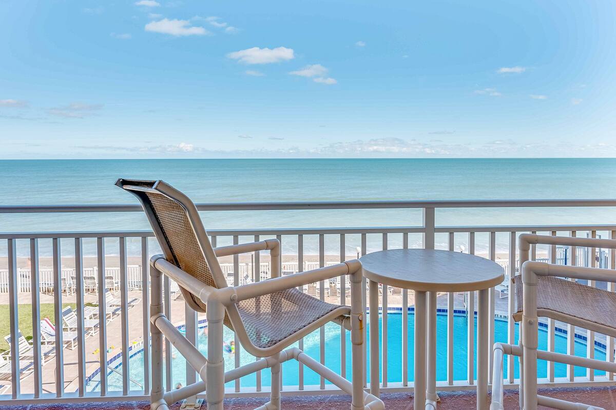 A balcony with a view of the ocean, a pool, and beach chairs.