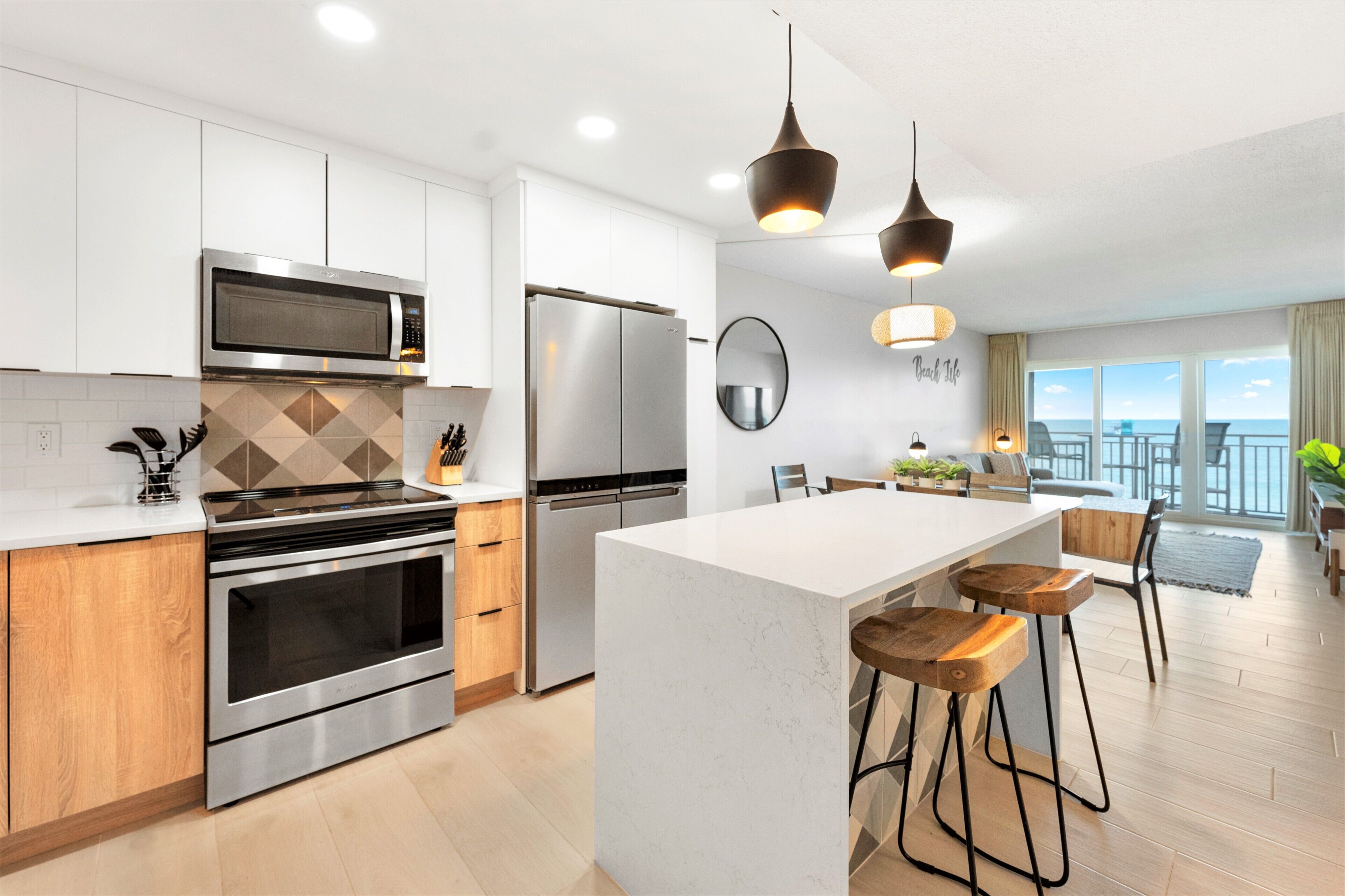 A modern kitchen with stainless steel appliances, a central island with stools, and a view of the sea through large windows.