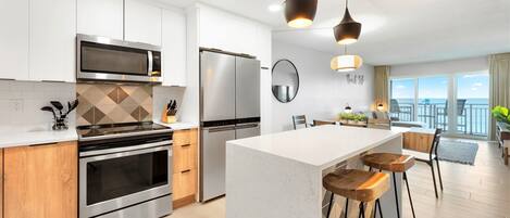 A modern kitchen with stainless steel appliances, a central island with stools, and a view of the sea through large windows.