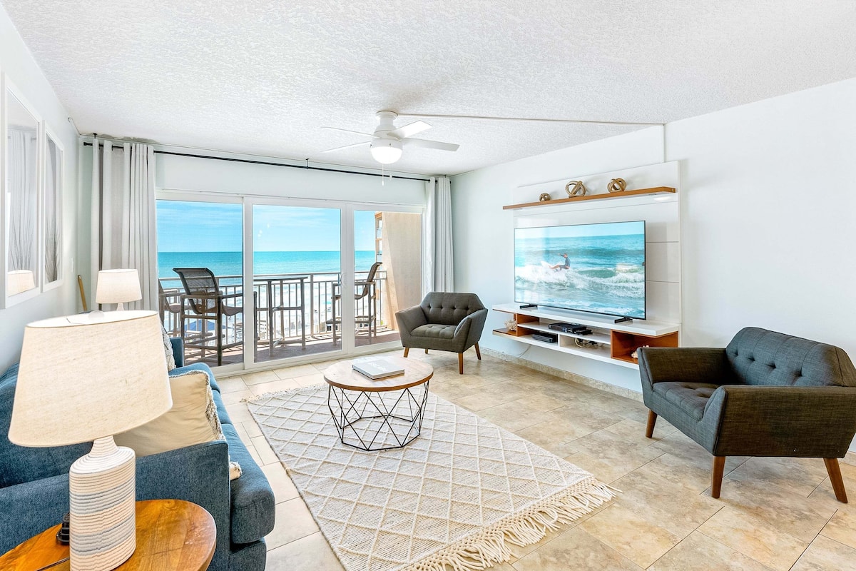 A living room with a TV, two sofas, a coffee table, and a view of the ocean.