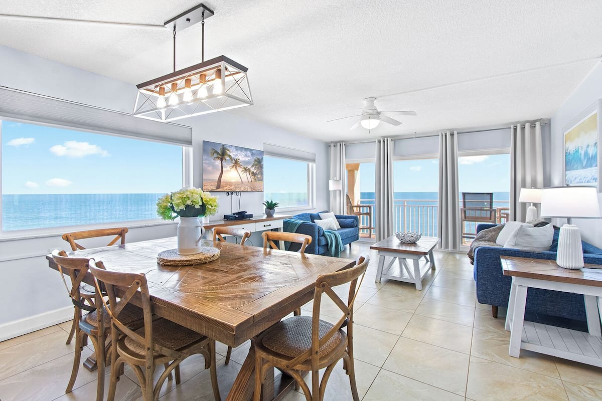 A spacious living area with a dining table, chairs, a sofa, and a balcony with a view of the ocean.