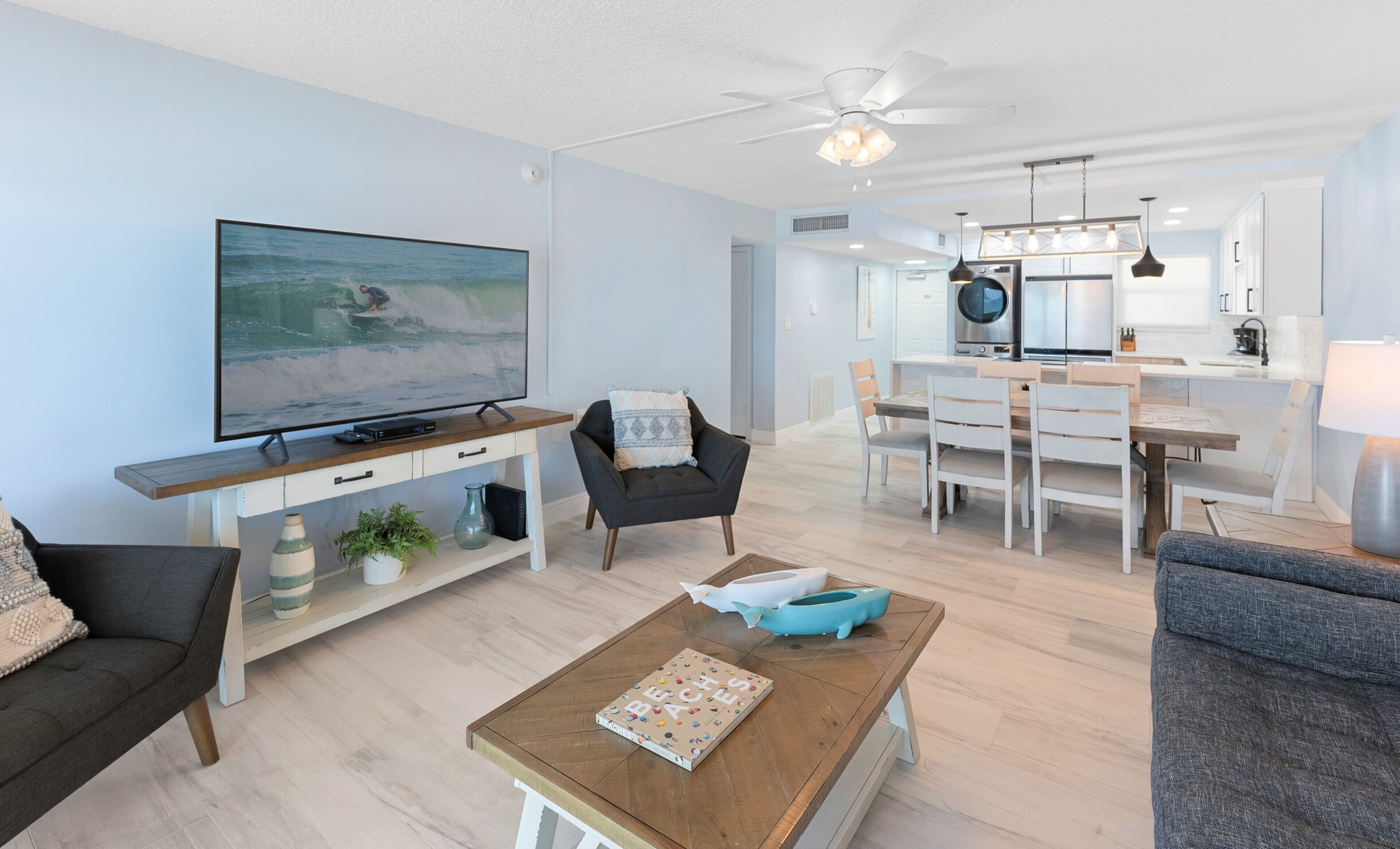 A modern living room with a flat-screen TV, a dining area with a wooden table and chairs, and a kitchen with stainless steel appliances.