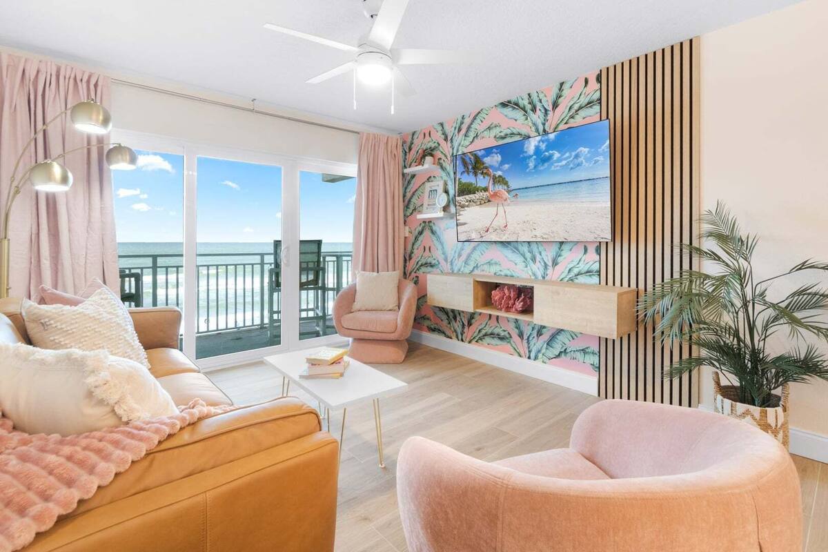 A living room with a sofa, armchair, and coffee table, a TV mounted on the wall, and a balcony with a view of the sea.
