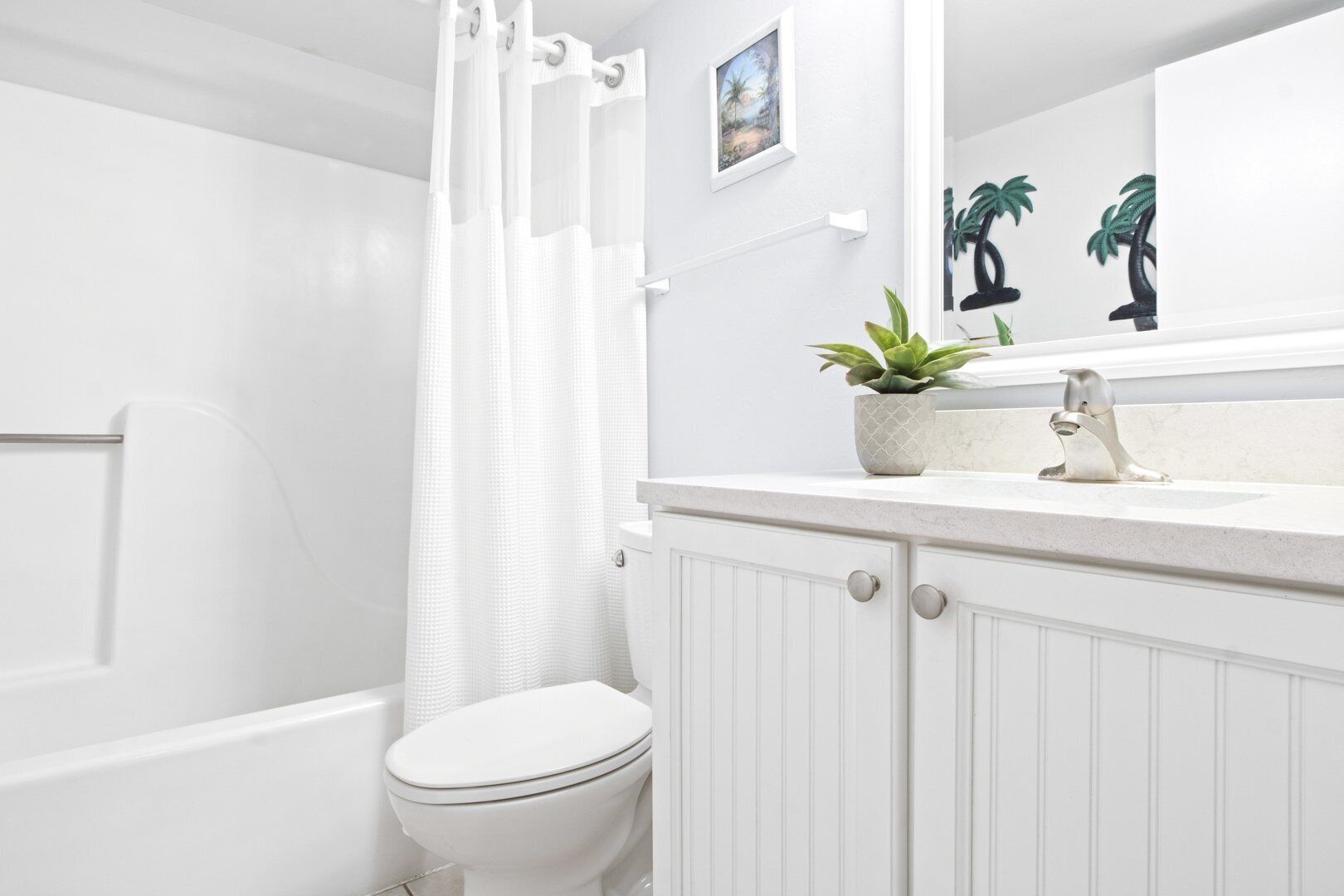 Une salle de bain blanche avec des toilettes, un rideau de douche, un lavabo agrémenté d’une plante et un miroir avec un tableau.