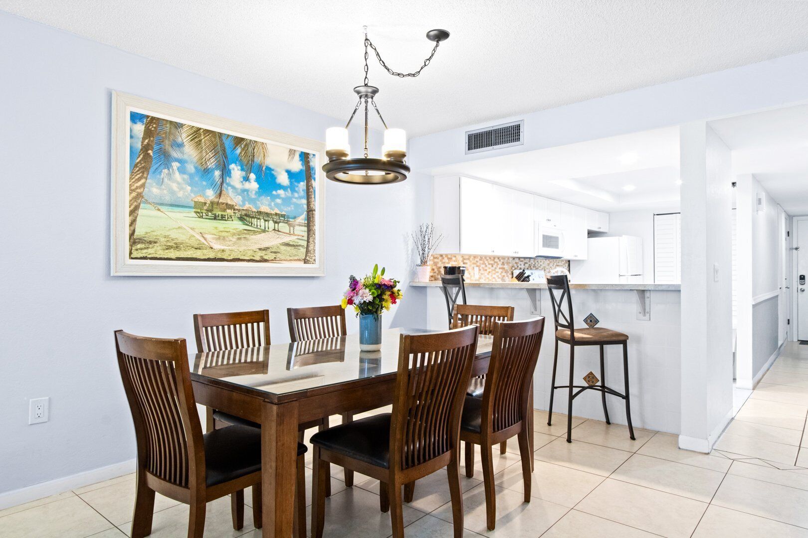 A dining area with a wooden table and chairs, a chandelier, a painting of a beach scene, and a kitchen in the background.