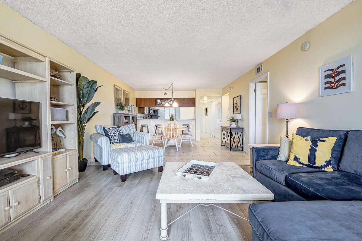 A spacious living area with a sofa, armchair, and a coffee table. There is a dining table with chairs, a kitchen area, and a large plant.