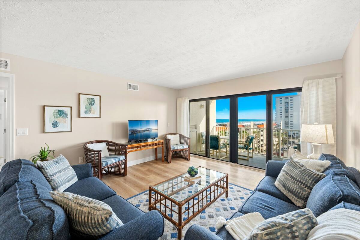 A living room with a sofa, coffee table, wicker chairs, a TV, and a balcony with a city view.