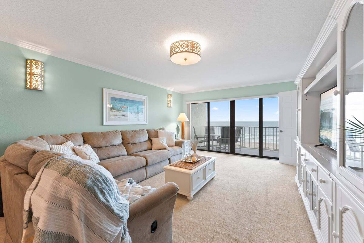 A living room with a sofa, a coffee table, a lamp, and a TV. There is a balcony with a view of the ocean.