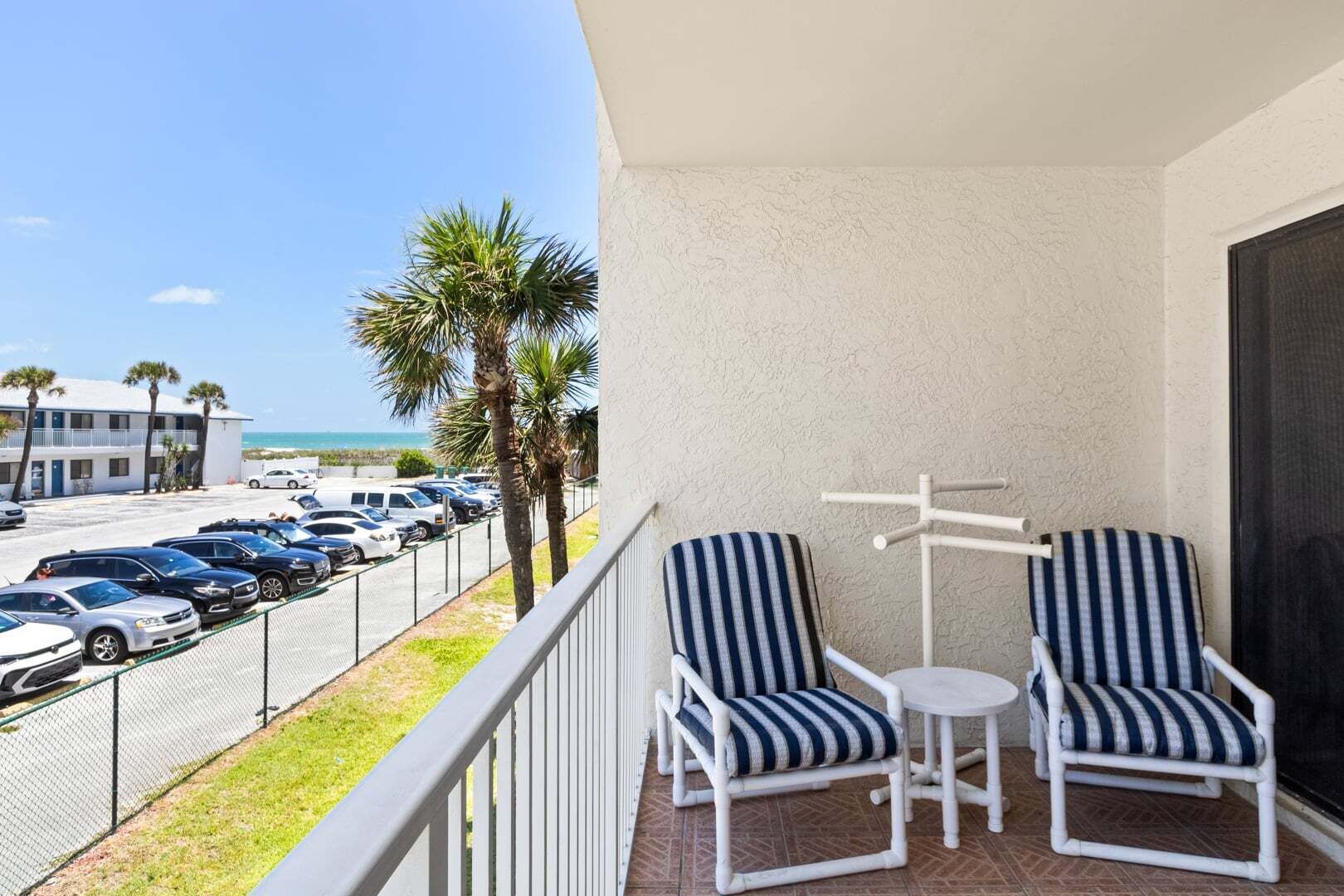 Een balkon met twee gestreepte stoelen en een kleine tafel, met uitzicht op een parkeerplaats en strand.