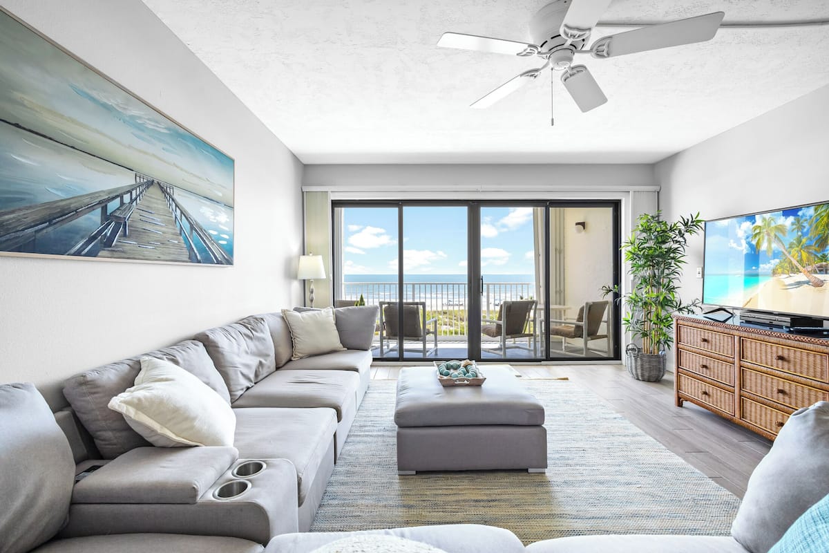 A modern living room with a sofa, ottoman, and television, a ceiling fan, and a view through a glass door to the outdoors.