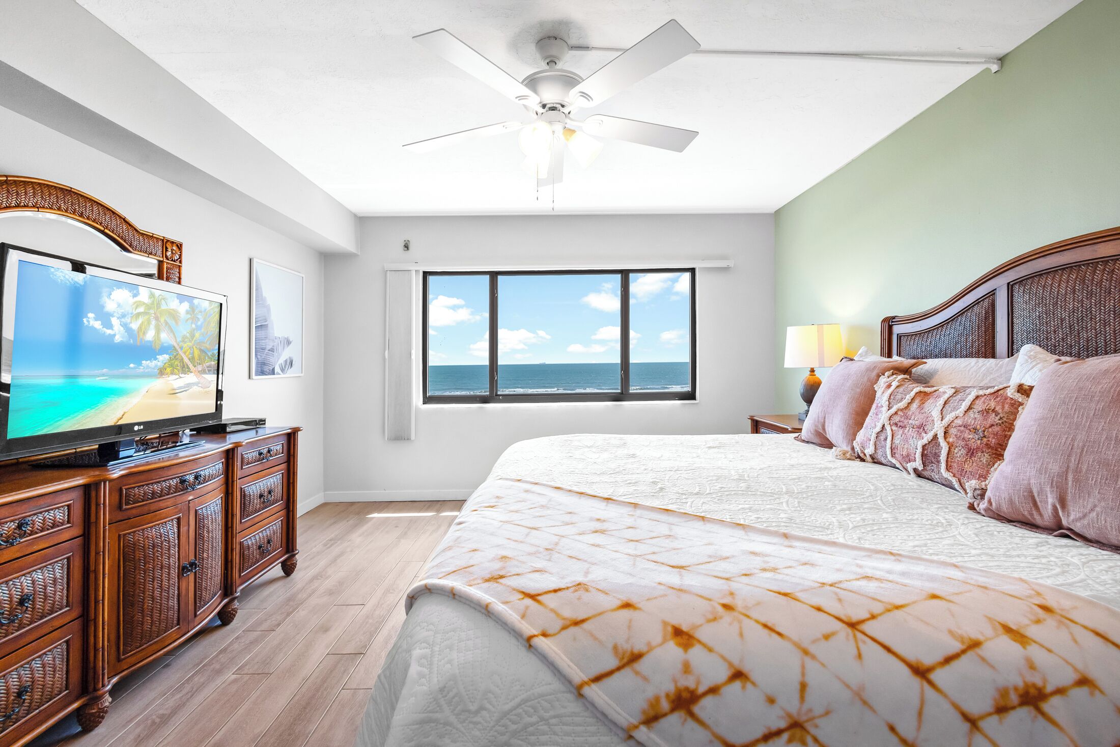 Un dormitorio con una cama grande, un televisor sobre un mueble de madera, un ventilador de techo y una ventana con vista al océano.