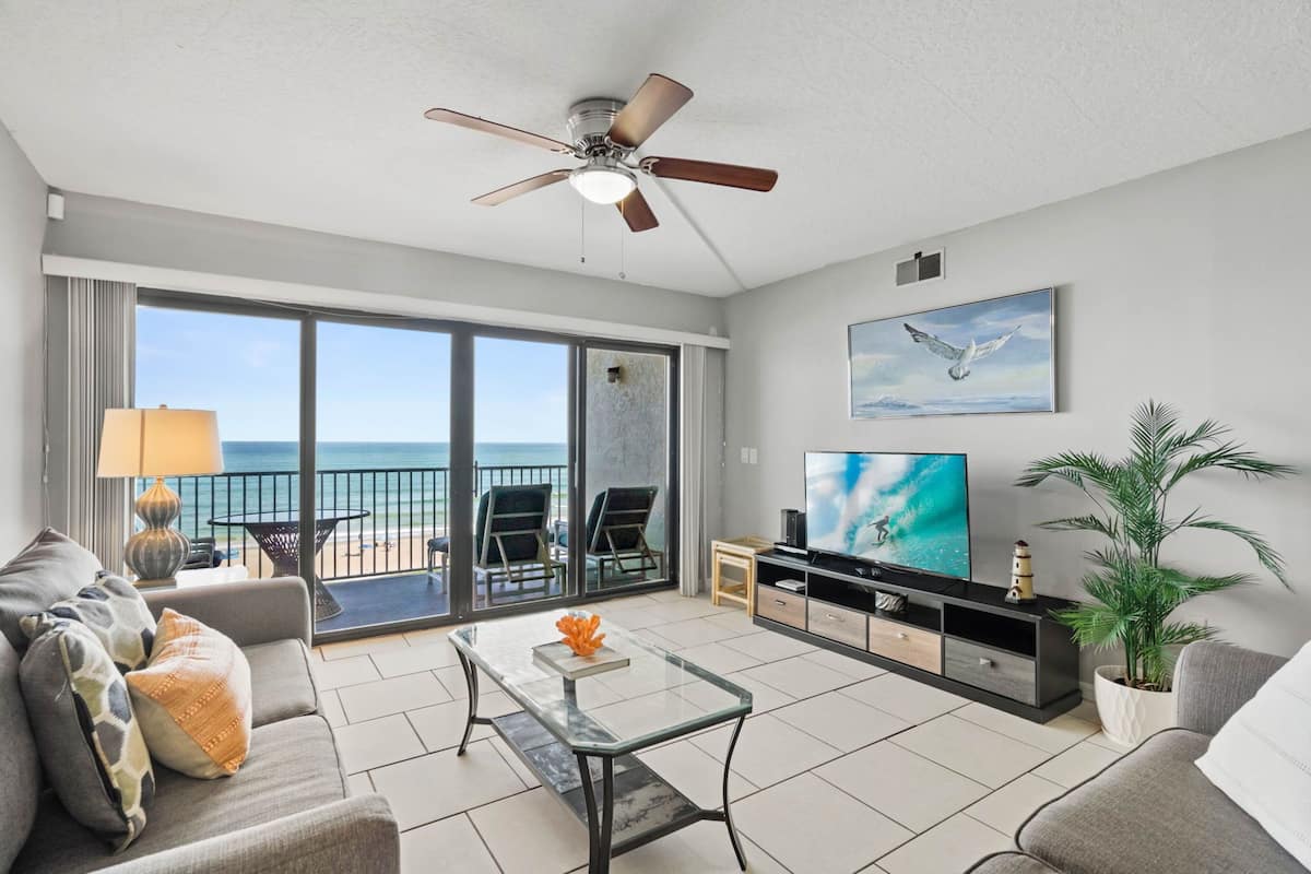 A modern living room with a balcony, a ceiling fan, a TV, and a view of the ocean.