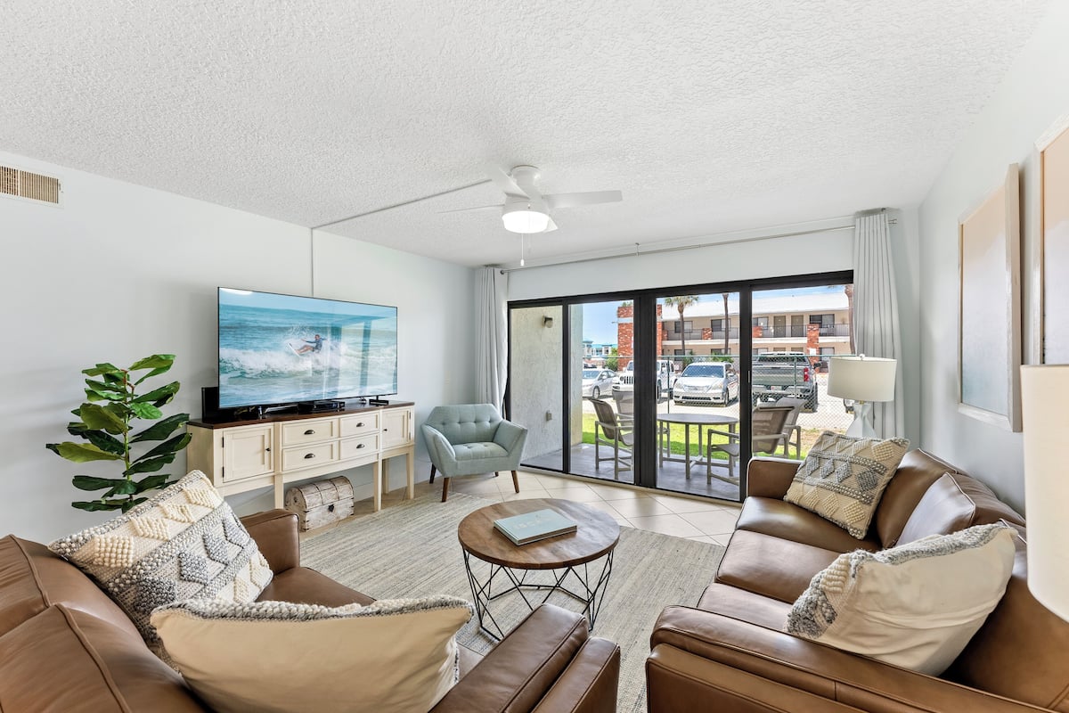 A living room with a television, two sofas, a coffee table, a ceiling fan, and a view of the outside.