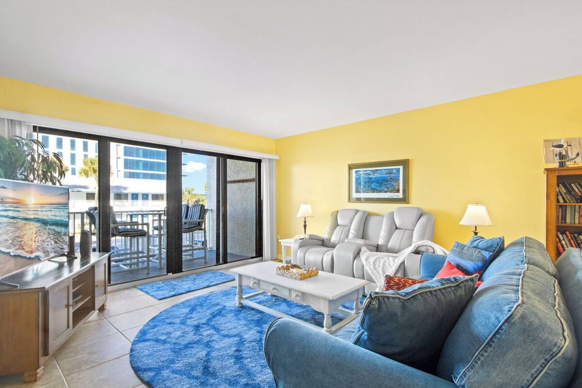 A living room with a sofa, armchairs, a coffee table, and a TV. There is a sliding glass door leading to an outdoor area with a pool and chairs.