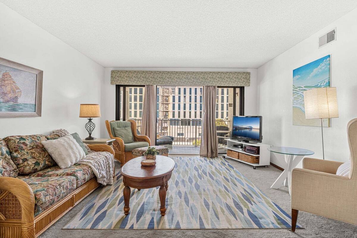 A living room with a sofa, armchair, coffee table, TV, and a balcony with curtains.