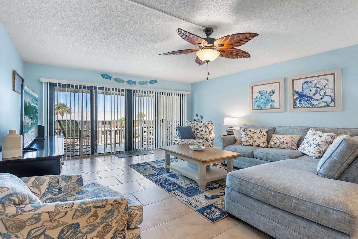 A living room with a ceiling fan, a sofa, a patterned armchair, a coffee table, and a view through a sliding glass door.