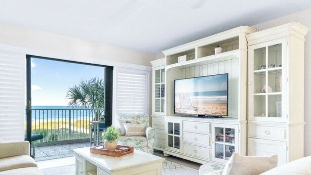 A spacious living room with a large TV, white cabinetry, and a balcony with a view of the beach. - Sandcastles - Cocoa Beach Oceanfront Condos with Heated Pool Tennis Pet-Friendly (Cocoa Beach)