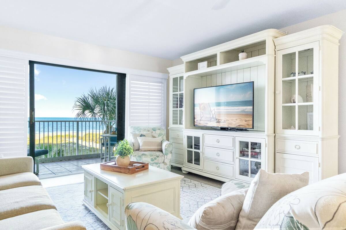 A spacious living room with a large TV, white cabinetry, and a balcony with a view of the beach.