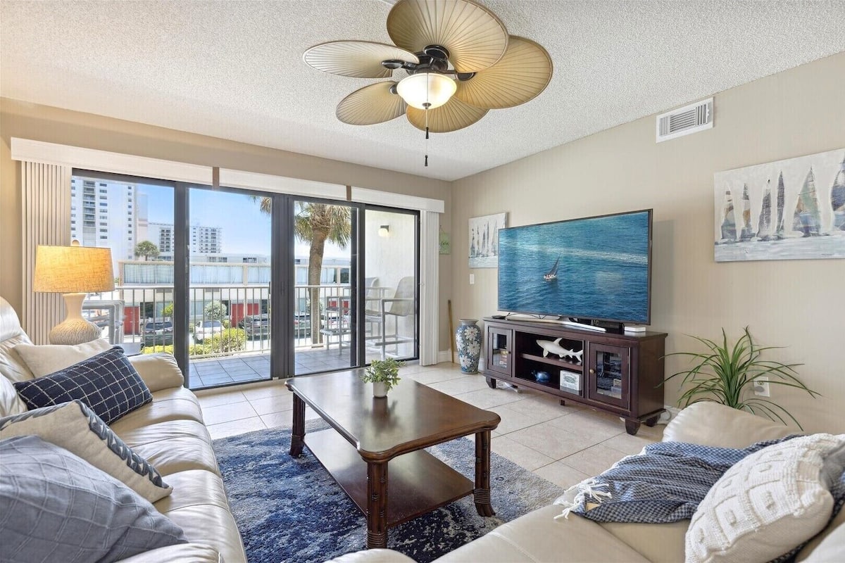 A spacious living room with a large TV, comfortable seating, a ceiling fan, and a balcony with a city view.