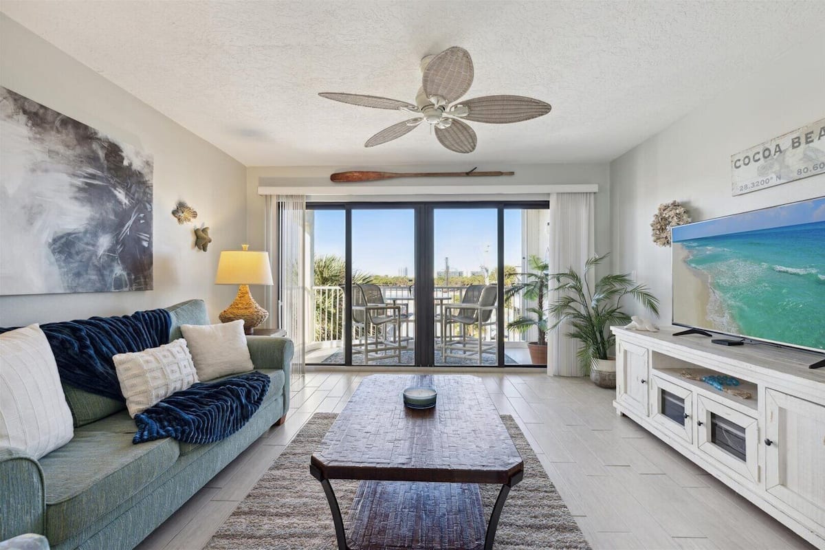 A living room with a sofa, coffee table, TV, ceiling fan, and a view of the outdoors through a sliding door.