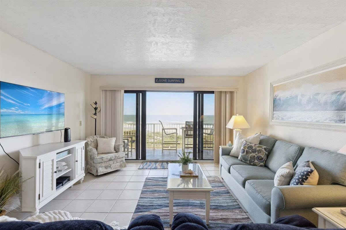 A living room with a television, a sofa, a coffee table, and a balcony with a view.