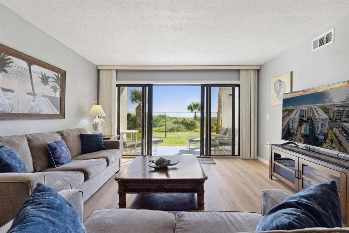 A living room with a sofa, coffee table, TV, and a view of a pool and greenery.