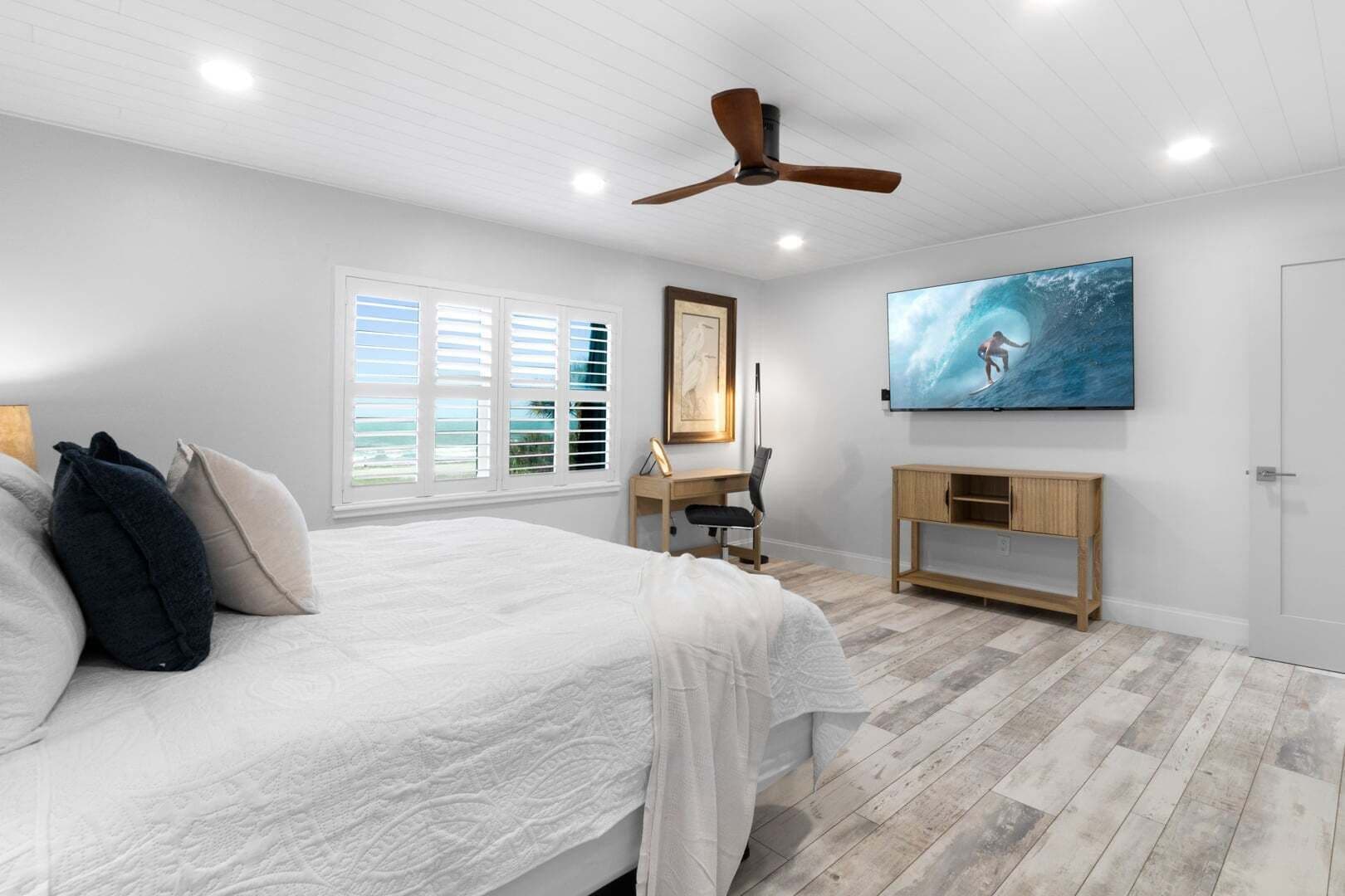 A bedroom with a bed, a desk, a chair, a ceiling fan, a TV, and a window with blinds.