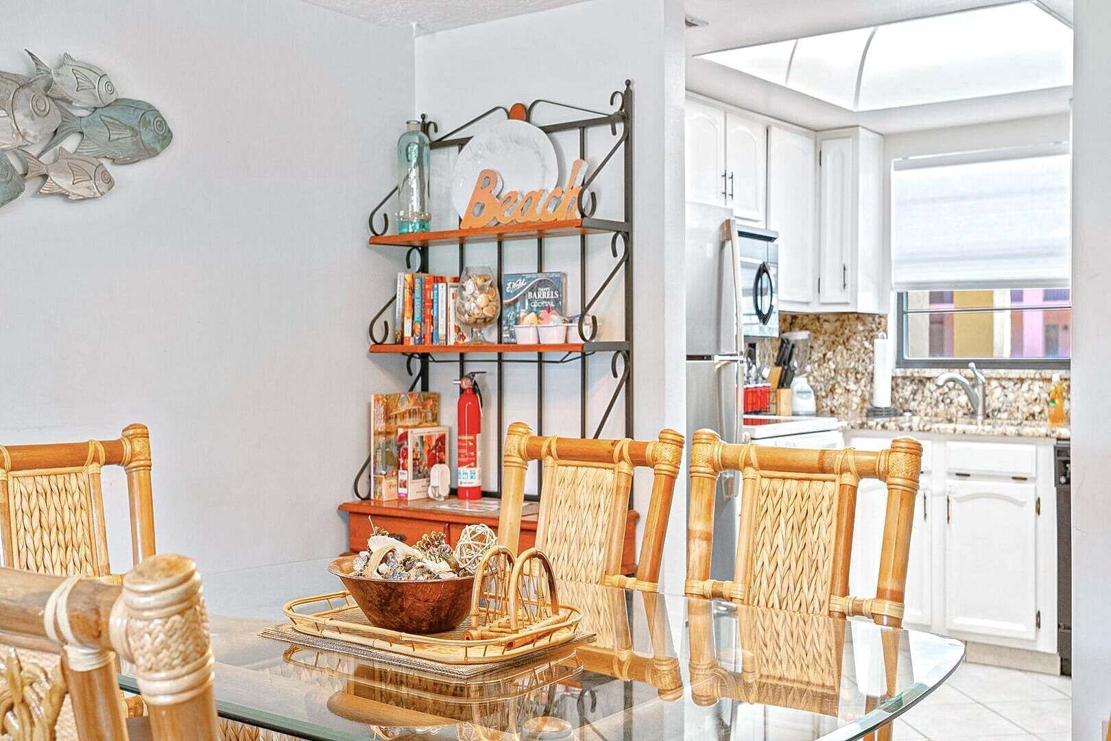 A dining area with a glass table, wicker chairs, a metal shelf with books and decorative items, and a view of the kitchen in the background.