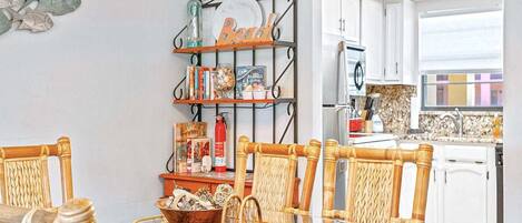 A dining area with a glass table, wicker chairs, a metal shelf with books and decorative items, and a view of the kitchen in the background.