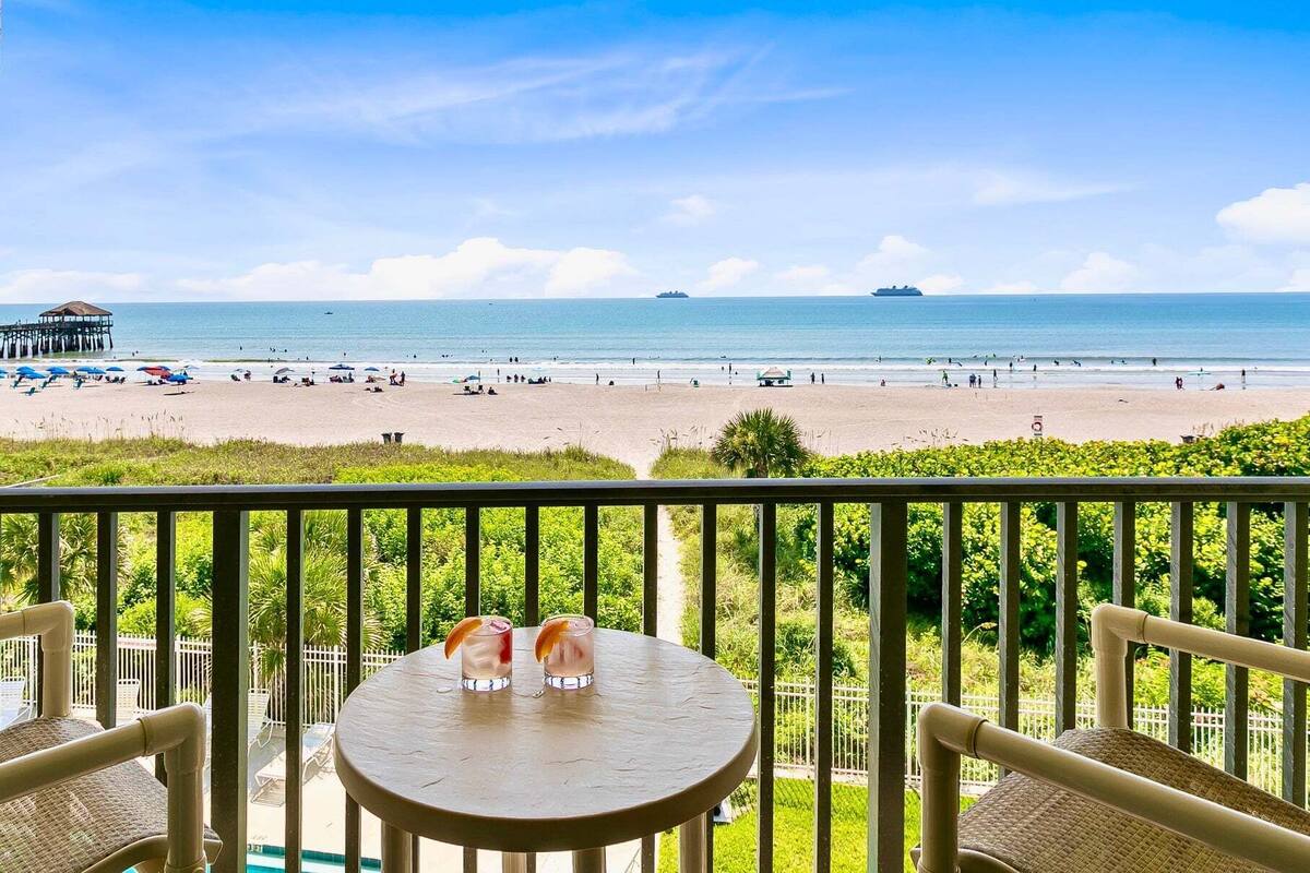 Un balcone con tavolo e sedie che si affaccia su una spiaggia con ombrelloni e un molo.