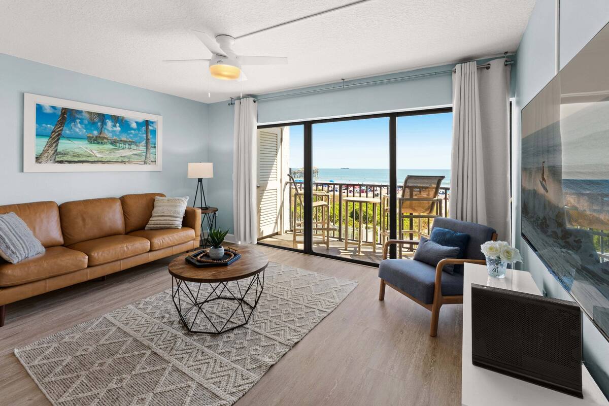 Un soggiorno con un divano in pelle marrone, un tavolino rotondo, un quadro che raffigura una spiaggia e un balcone con vista sull'oceano.