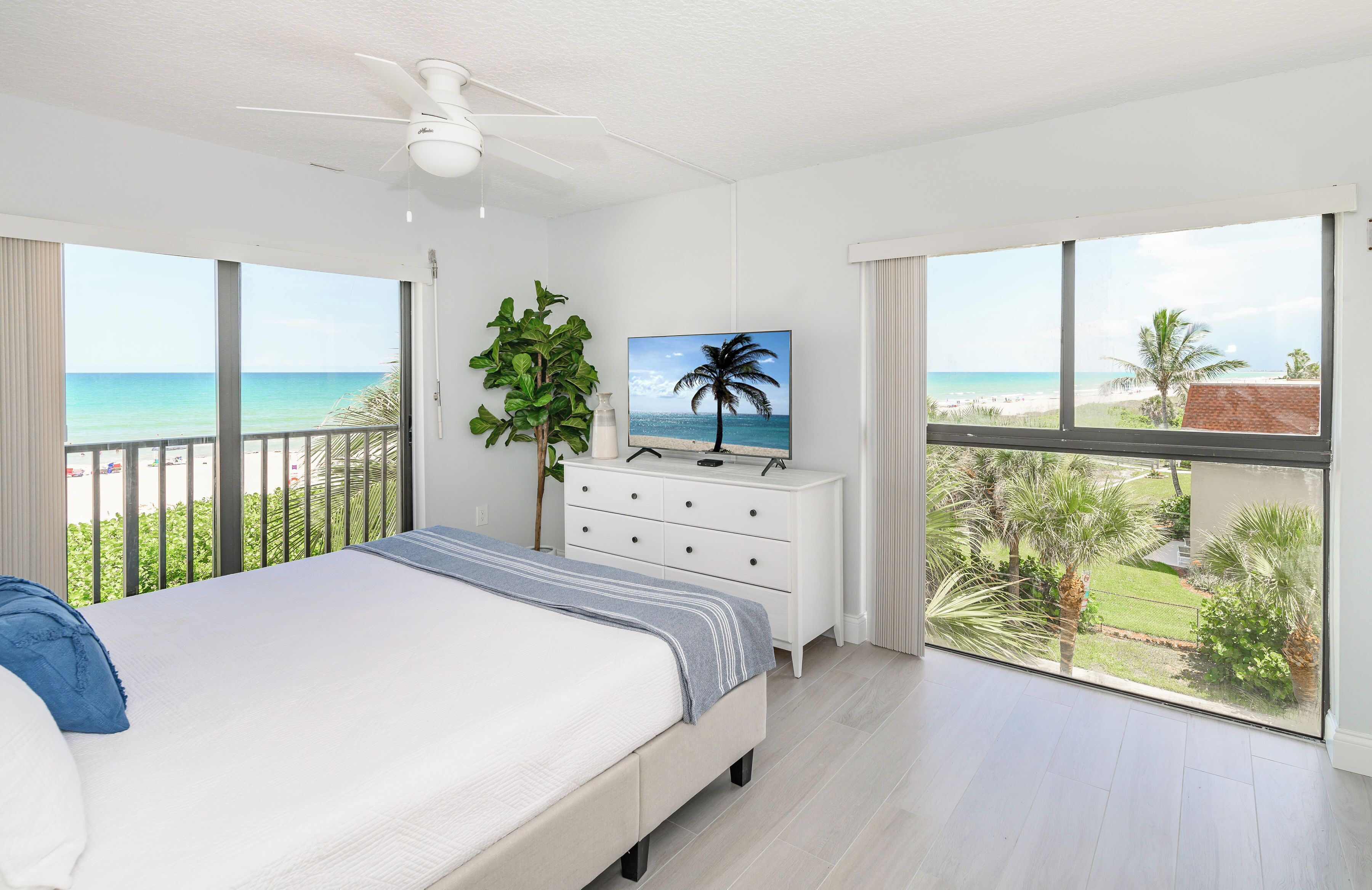 A bedroom with a bed, a dresser, a ceiling fan, a TV, and large windows with a view of the beach and palm trees.