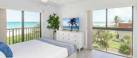 A bedroom with a bed, a dresser, a ceiling fan, a TV, and large windows with a view of the beach and palm trees.