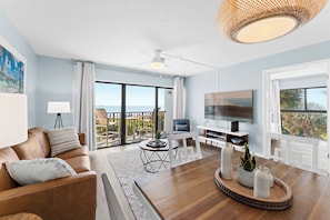 A modern living room with a sofa, TV, and dining area, large windows, and a balcony with a view. - Chateau by the Sea - Cocoa Beach Oceanfront Condos next to the Pier and Port Canaveral (Cocoa Beach)