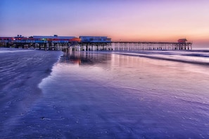 A pier extending into the water with buildings on stilts, a beach, and a sunset. - Chateau by the Sea - Cocoa Beach Oceanfront Condos next to the Pier and Port Canaveral (Cocoa Beach)