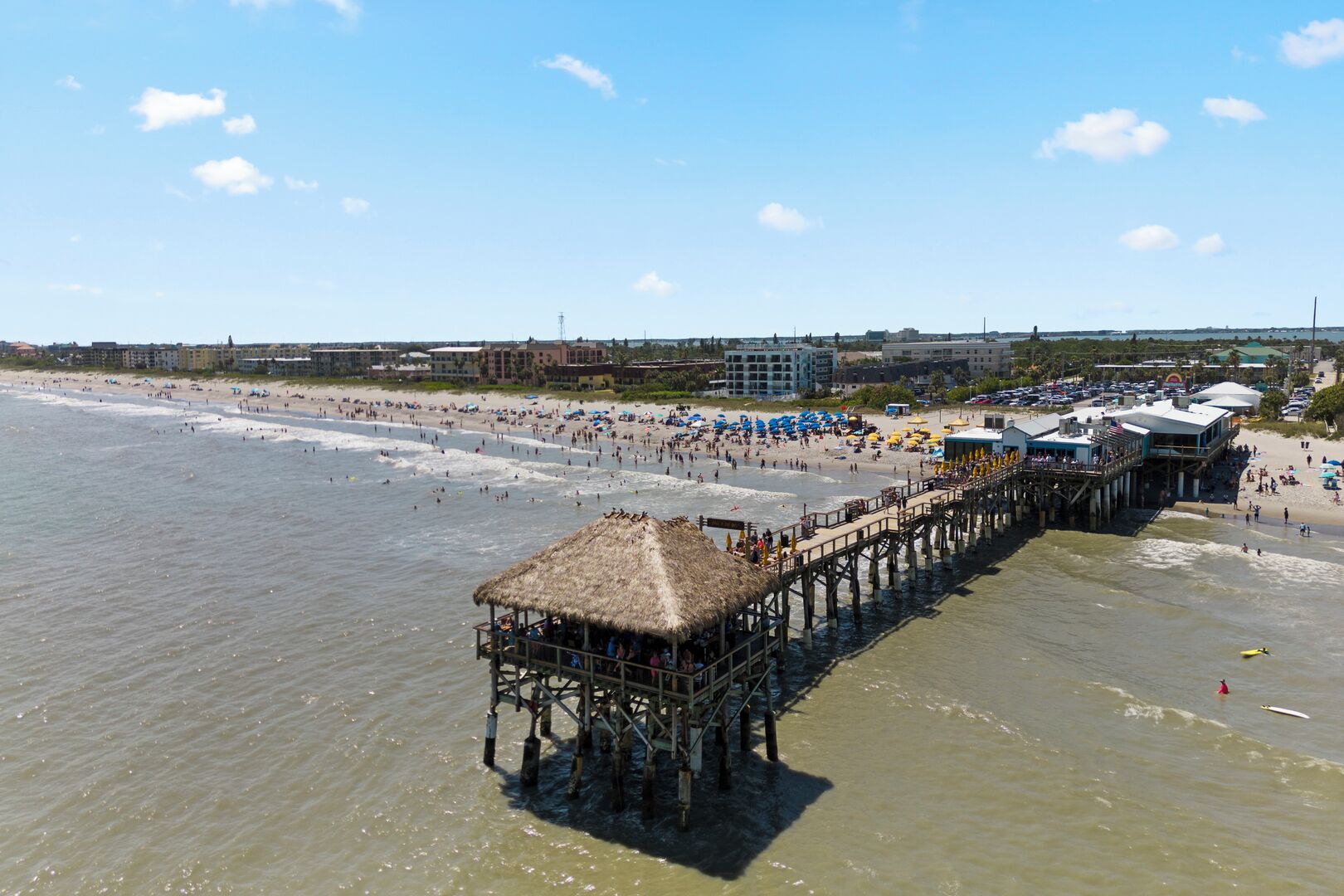 A pier extending into the sea with a thatched-roof structure at the end, surrounded by beachgoers and beach umbrellas.