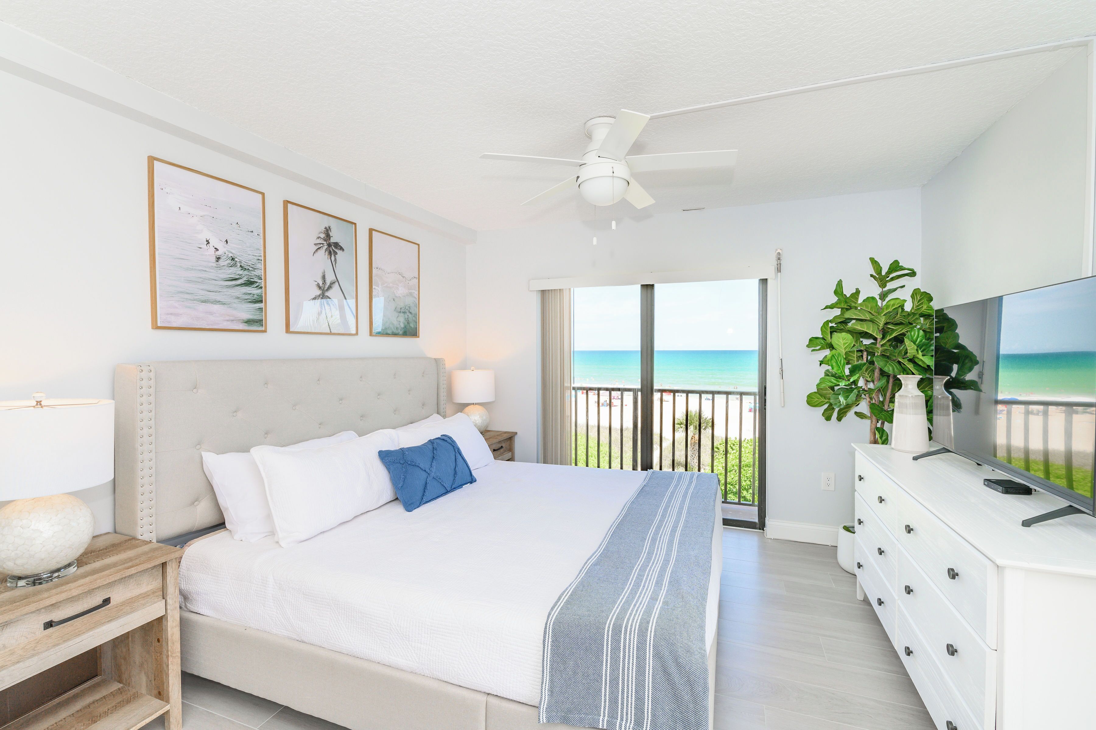 A bedroom with a bed, a nightstand, a dresser, a ceiling fan, and a balcony with a view of the ocean.