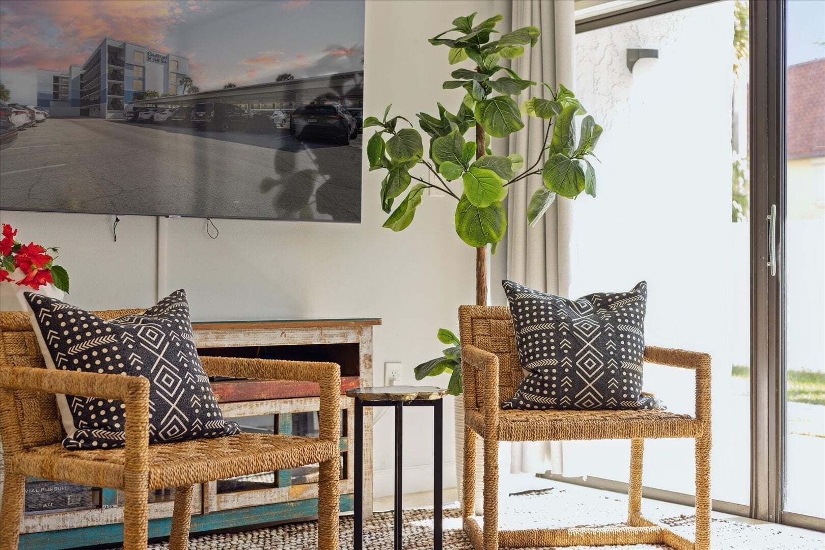 Des chaises en osier ornées de coussins à motifs, une plante en pot et une vue sur un bâtiment à travers une porte vitrée.