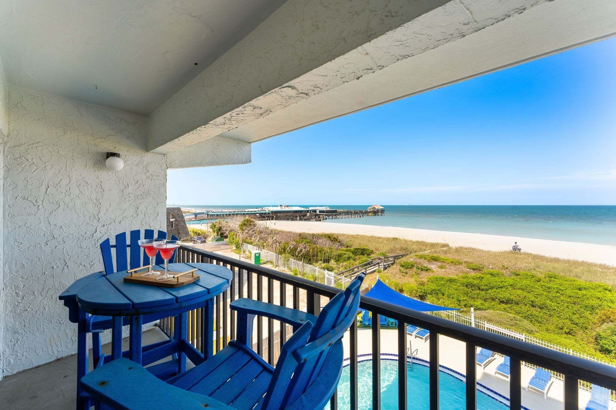 A balcony with blue chairs, a table, and a view of the beach and ocean.