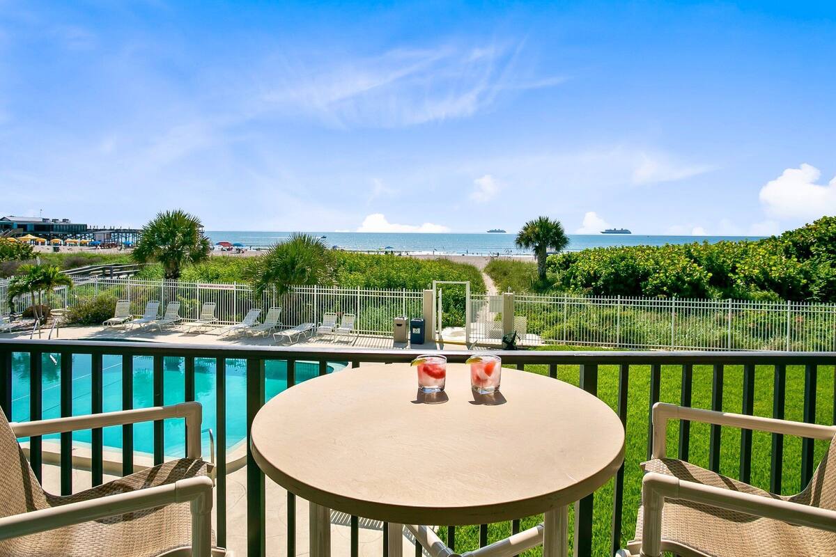Un balcone con vista sulla spiaggia, una piscina e due bevande su un tavolo.