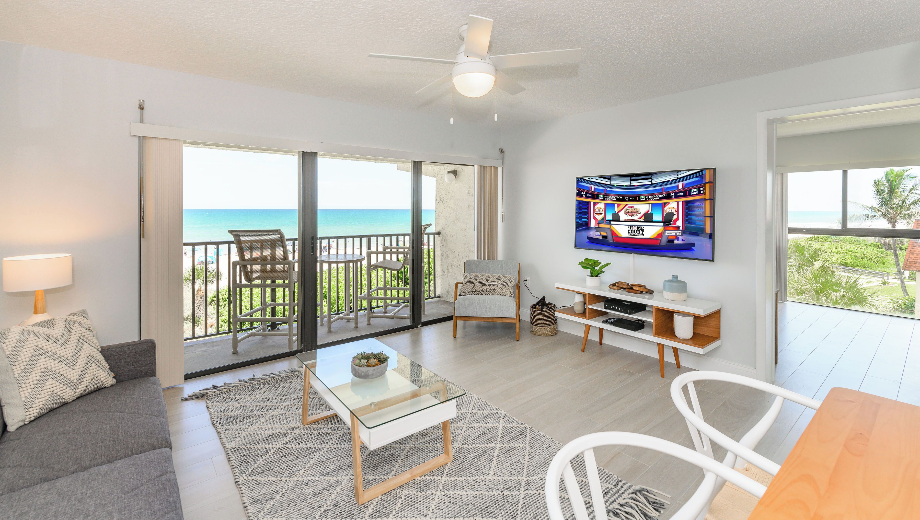 A living room with a TV, a sofa, a coffee table, and a balcony with a view of the ocean.