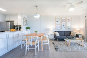 A modern kitchen with white cabinets, a dining area with a wooden table and white chairs, a gray sofa, and a ceiling fan. - Chateau by the Sea - Cocoa Beach Oceanfront Condos next to the Pier and Port Canaveral (Cocoa Beach)