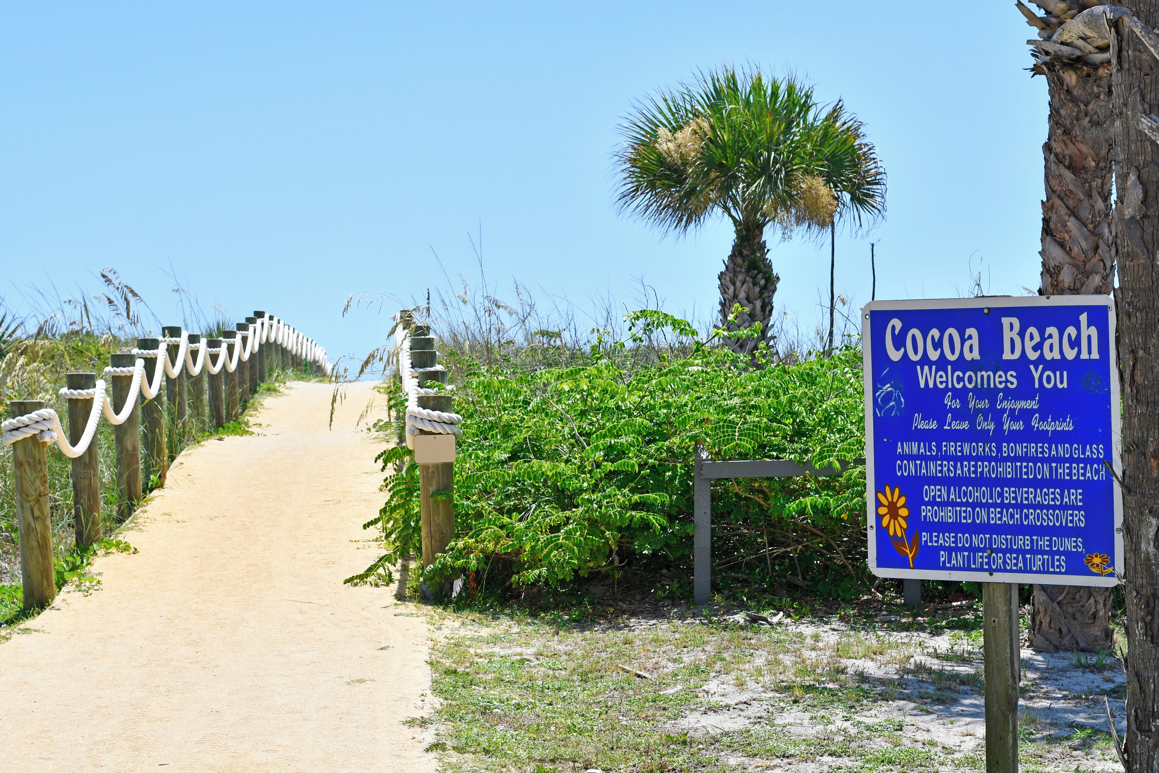 A beach path with a sign welcoming visitors to Cocoa Beach, prohibiting certain activities, and featuring a palm tree.