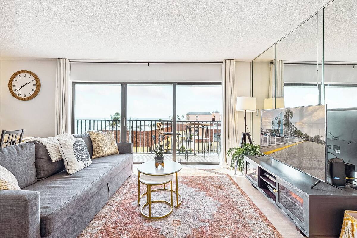 Un soggiorno moderno con un divano grigio, un tavolino rotondo, una televisione e un balcone con vista sugli edifici.