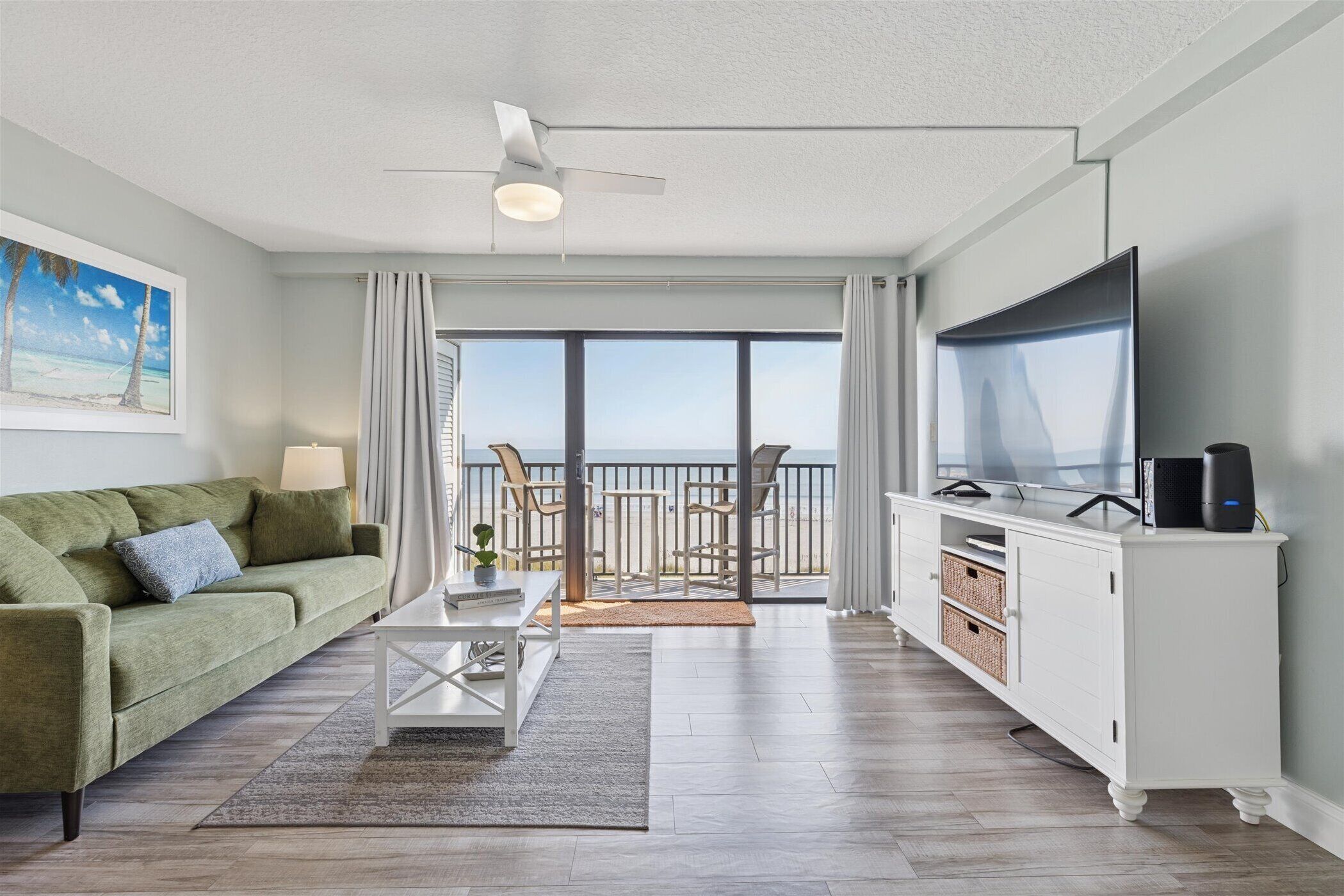 A modern living room with a green sofa, a glass coffee table, a flat-screen TV, and a balcony with a view of the ocean.