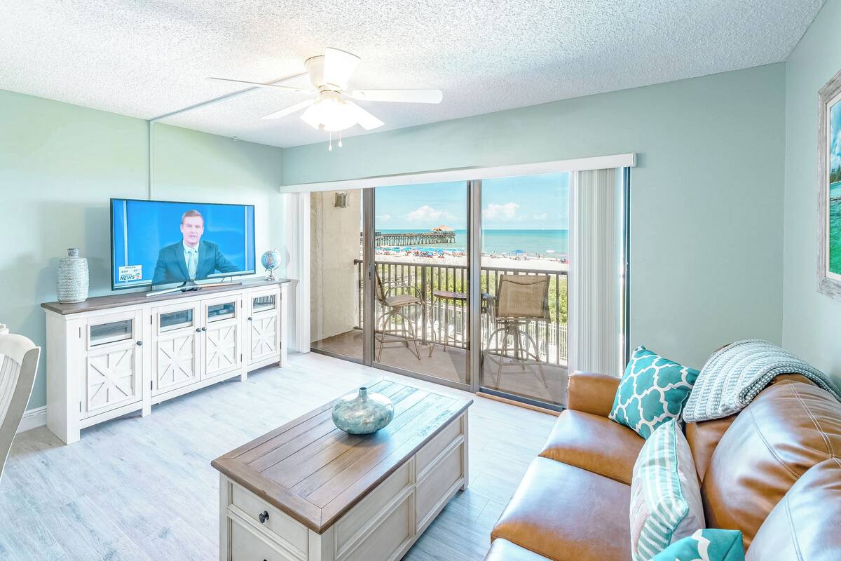 Un soggiorno con televisione, un tavolino in legno, una comoda poltrona in pelle e un balcone vista spiaggia e molo.
